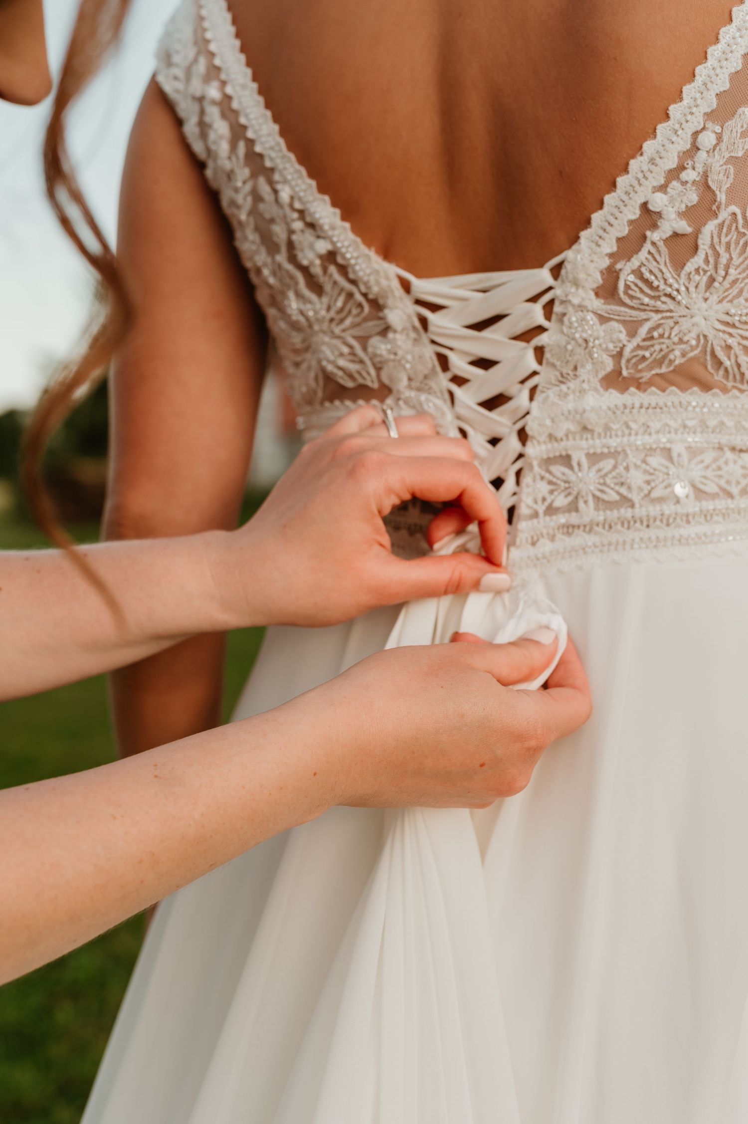 Bridesmaid fastening the lace corset back of a boho wedding dress with delicate floral embroidery details.