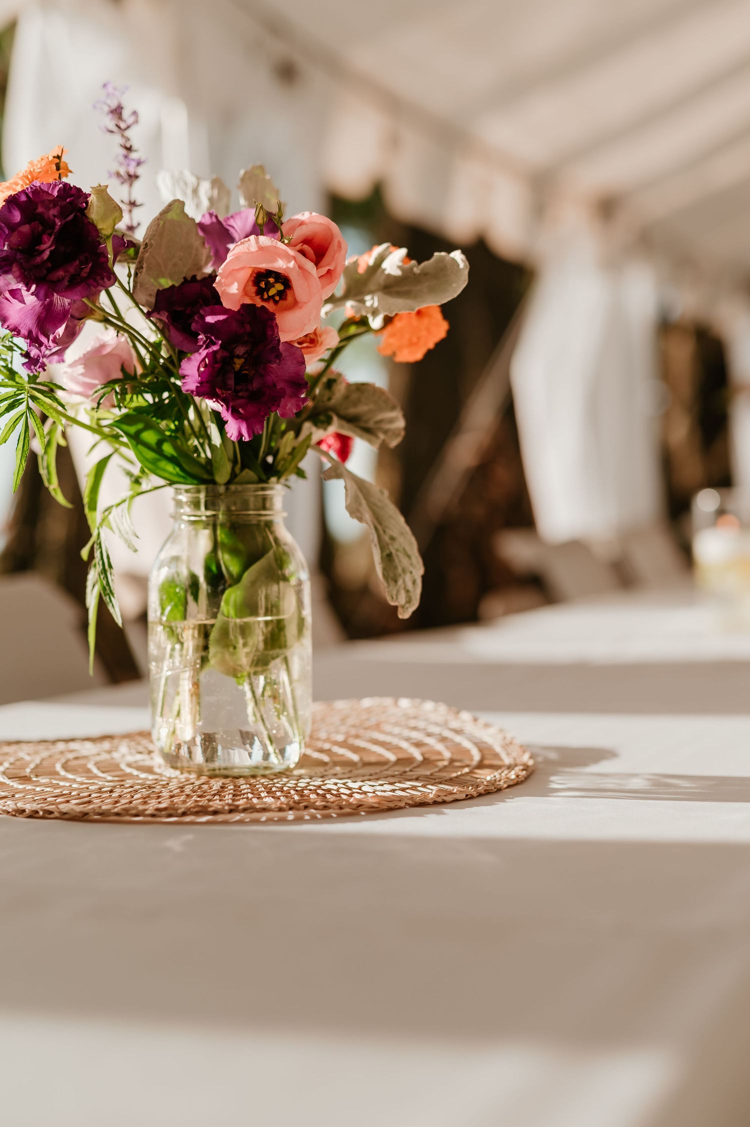 Mason jar with colorful wildflowers on woven placemat at elegant outdoor wedding reception table.