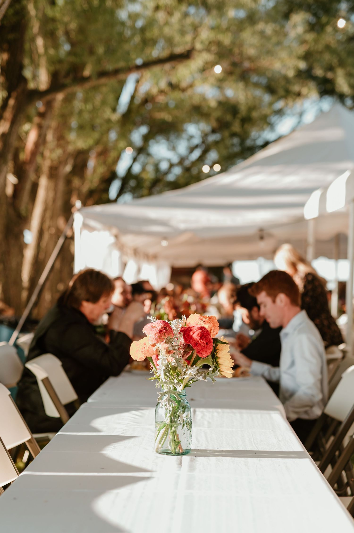Outdoor wedding reception with guests seated at white linen tables adorned with mason jar floral centerpieces under a canopy.