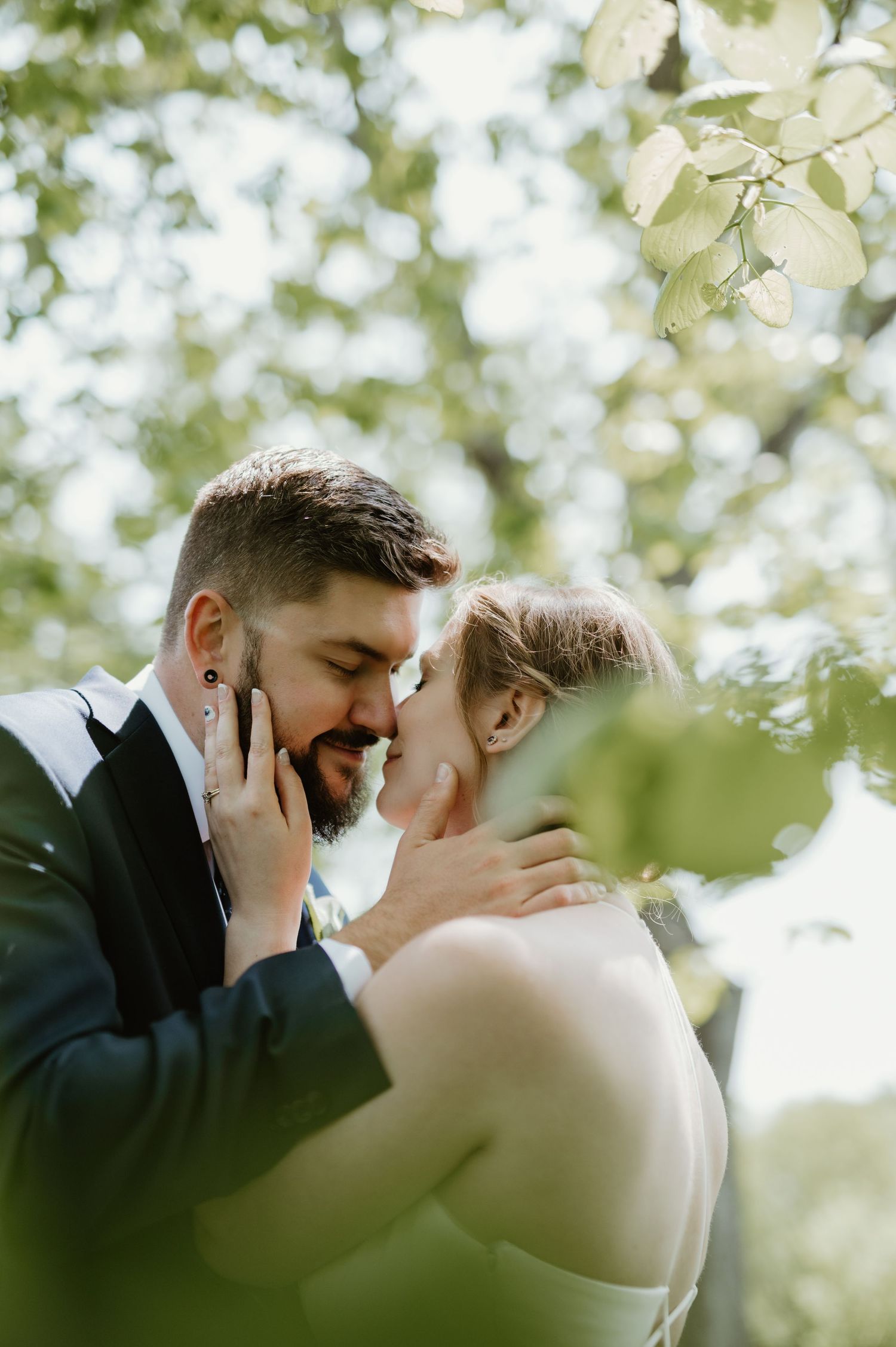 A bride and groom share a romantic almost-kiss under lush green trees in an outdoor wedding portrait.