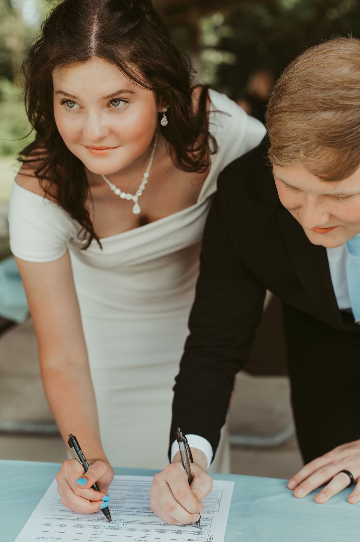 Bride and groom signing wedding certificate at elegant ceremony, bride wearing white dress and pearl necklace.