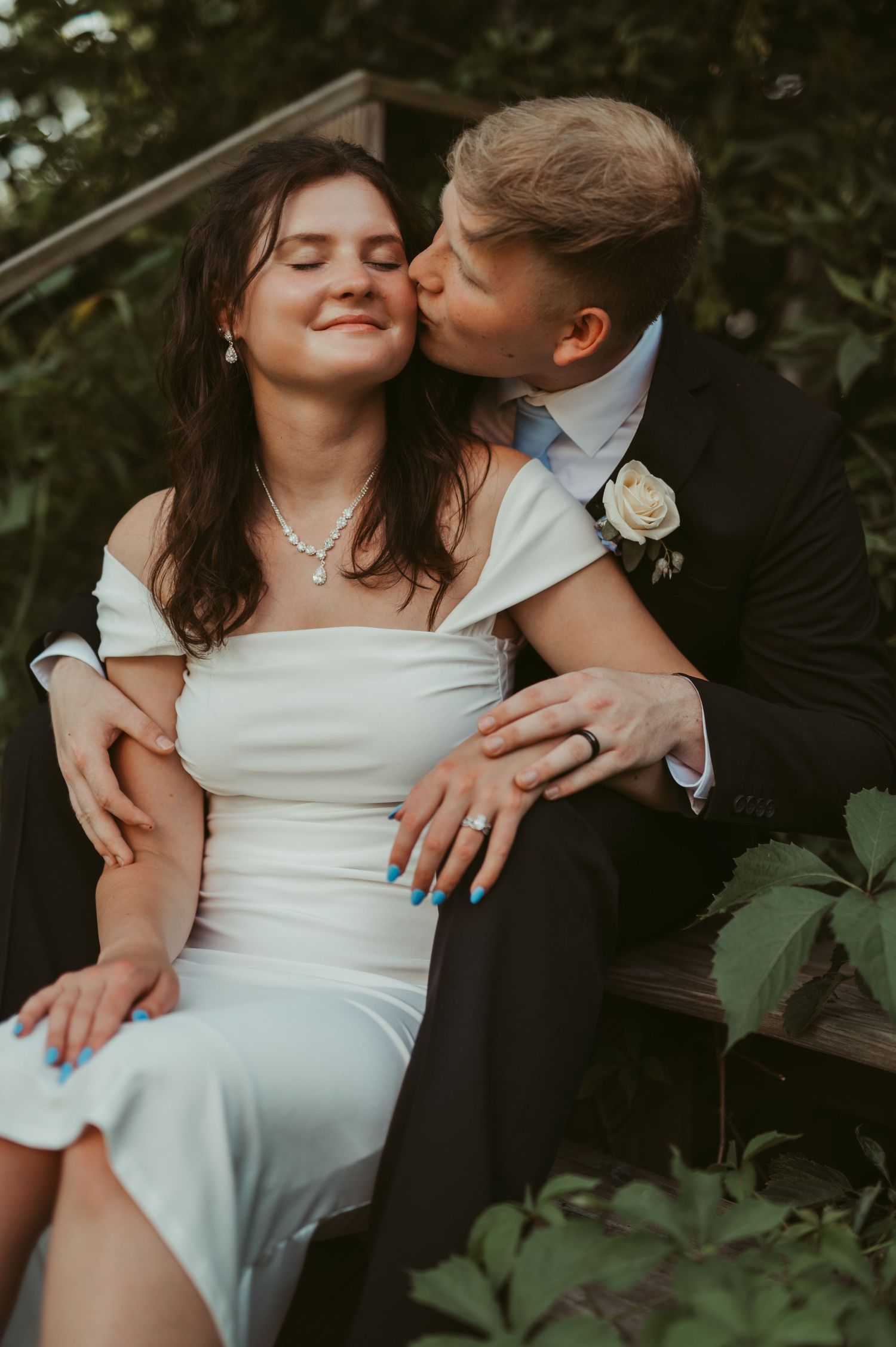 A bride in a white dress smiles as her groom in a black suit kisses her cheek while seated on steps outdoors.