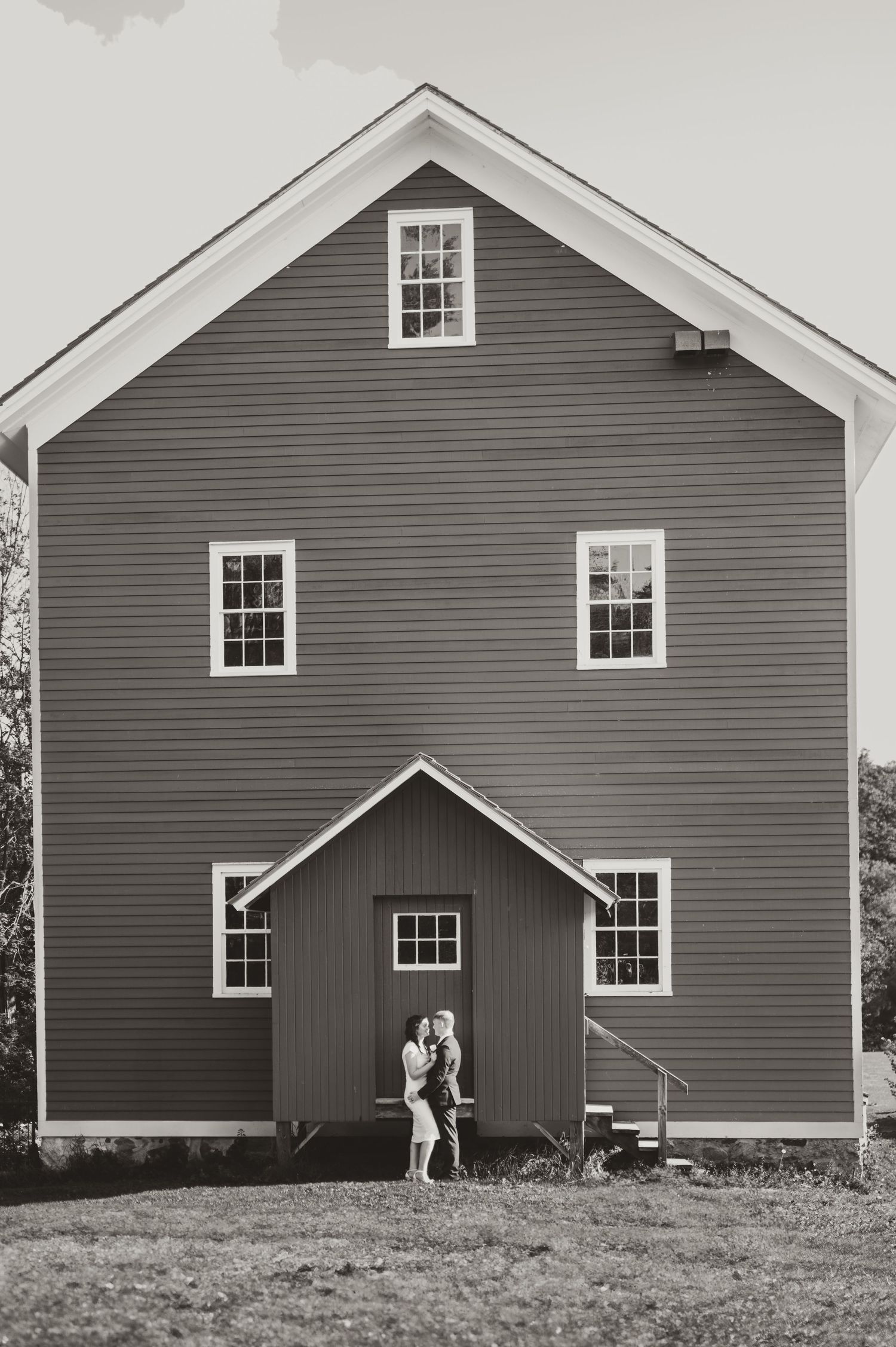 Black and white photo of a couple embracing in front of a large rustic barn with white-trimmed windows.