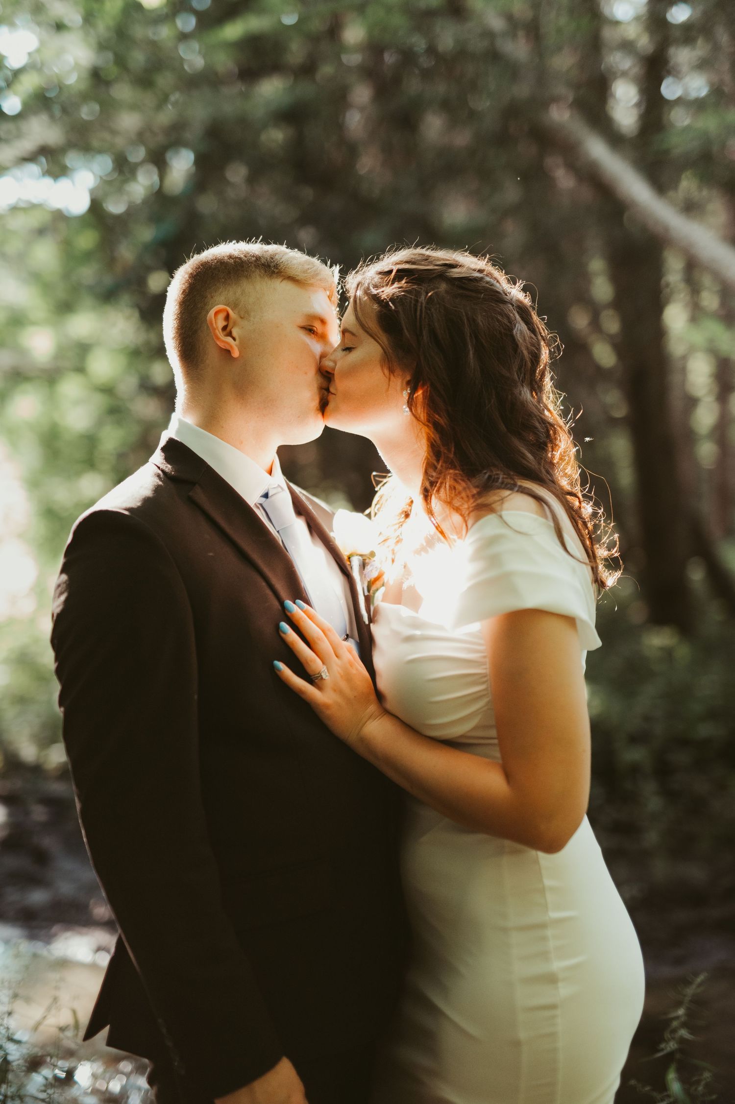 A couple shares a romantic kiss in a sunlit forest, dressed in wedding attire, a white gown and dark suit.