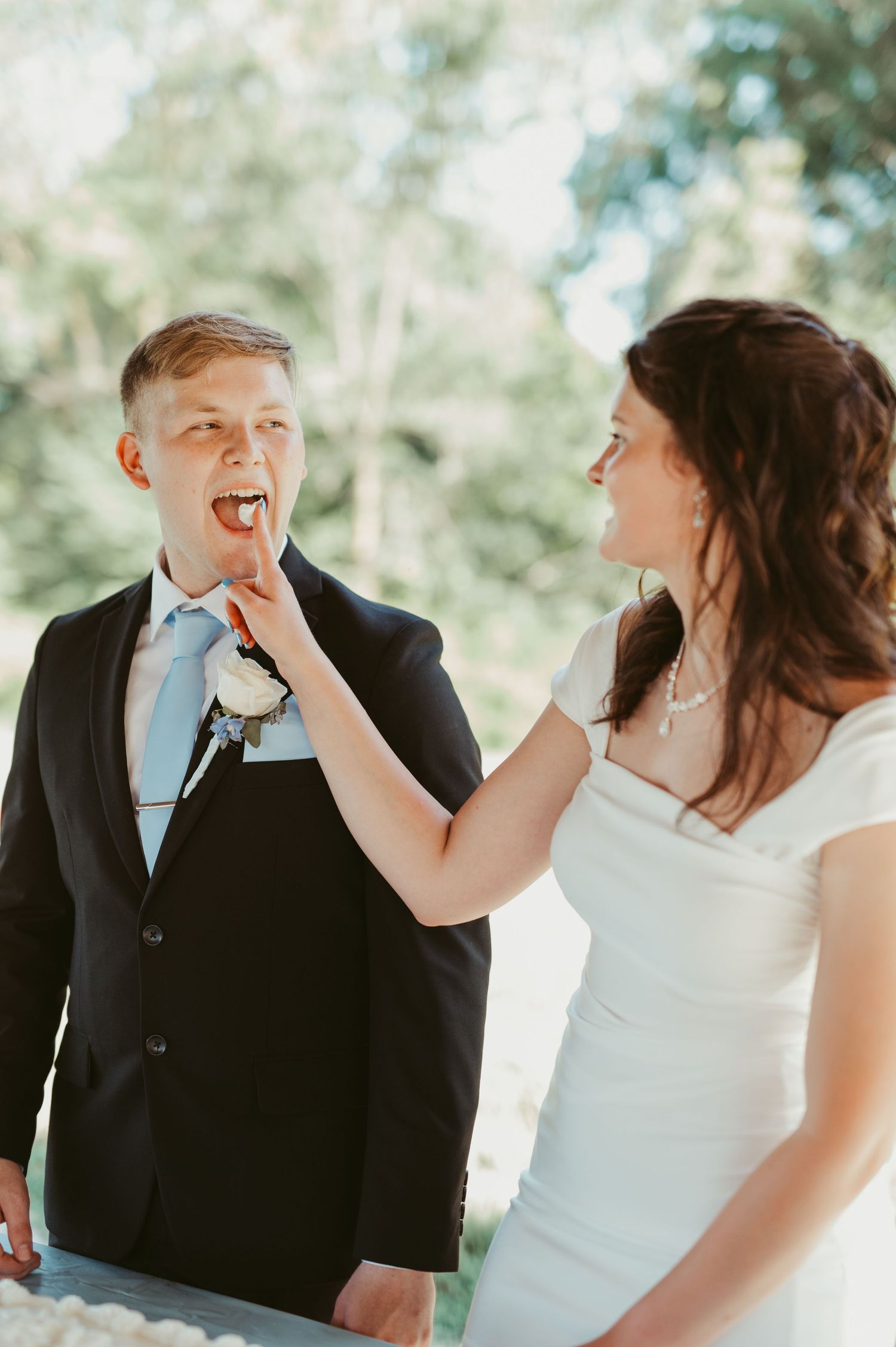 Bride playfully feeds wedding cake to groom in suit with blue tie and boutonniere outdoors.