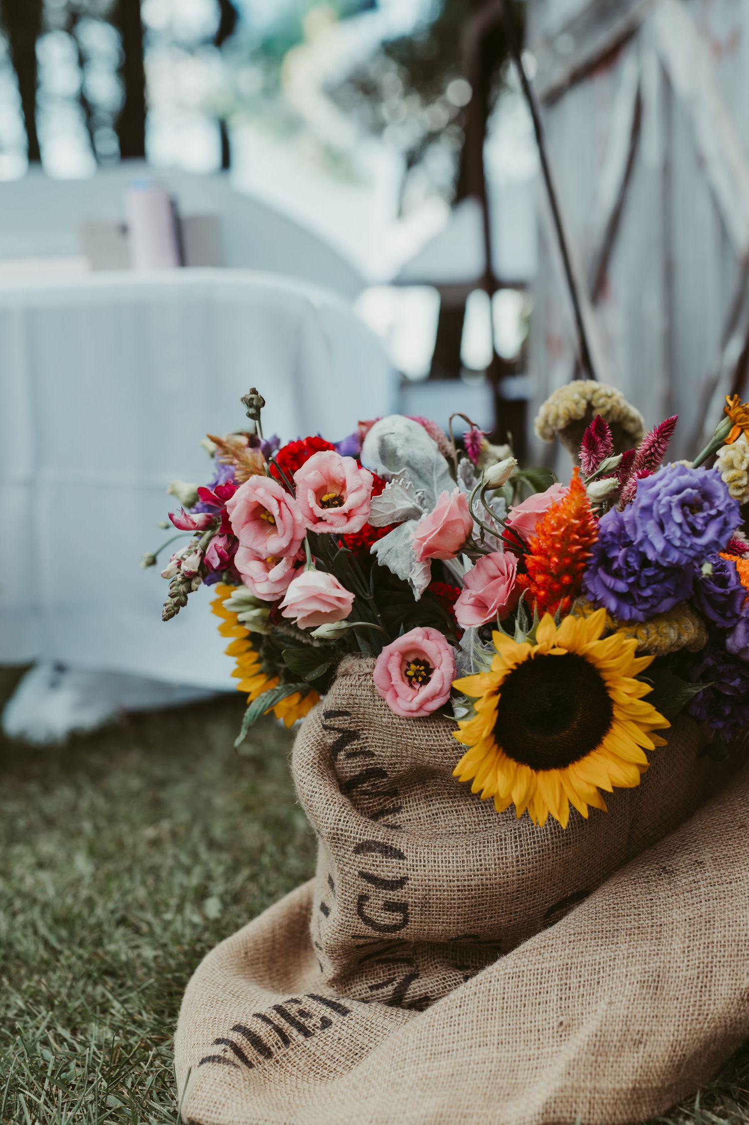 Colorful wildflower bouquet with sunflowers in a rustic burlap sack at an outdoor wedding reception.