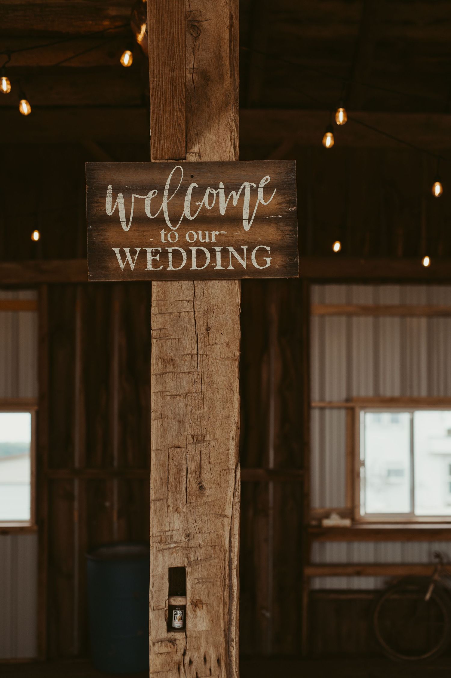 Rustic wooden welcome to our wedding sign mounted on a barn post with warm string lights in the background.