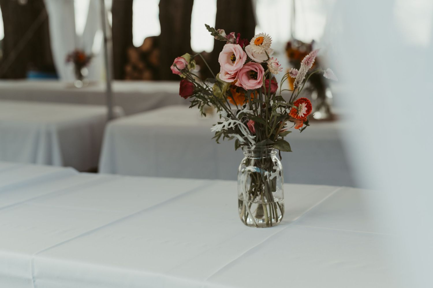 A glass jar vase with pink, red, and orange flowers as a centerpiece on a white table at a wedding reception.