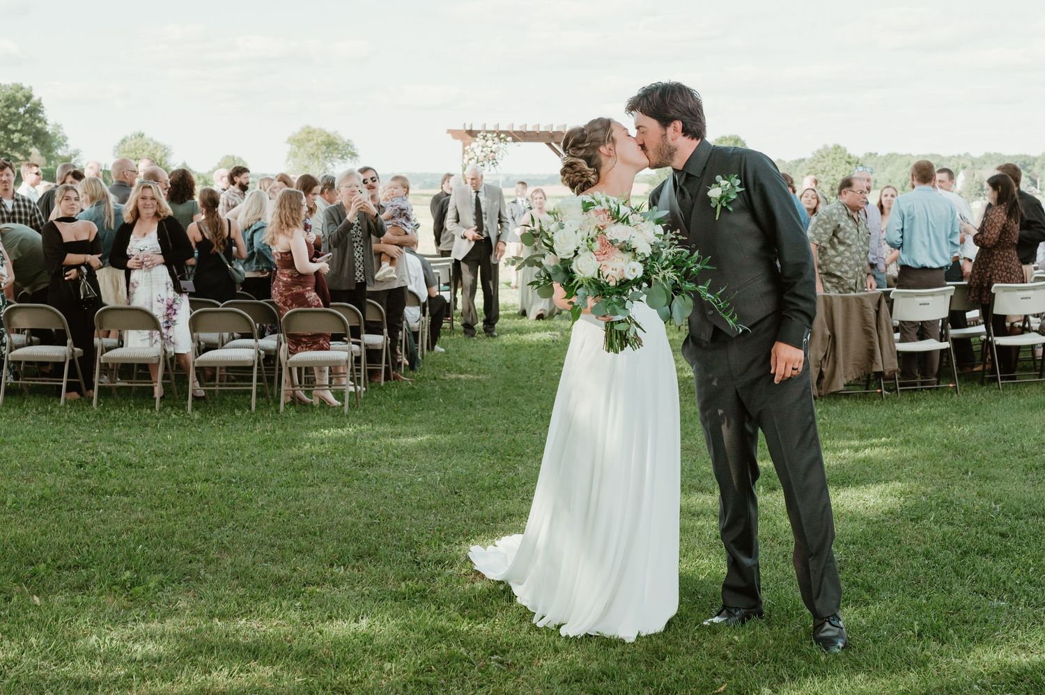 Bride and groom share first kiss on green lawn at outdoor wedding ceremony with guests seated in background in Jackson, Wisconsin.
