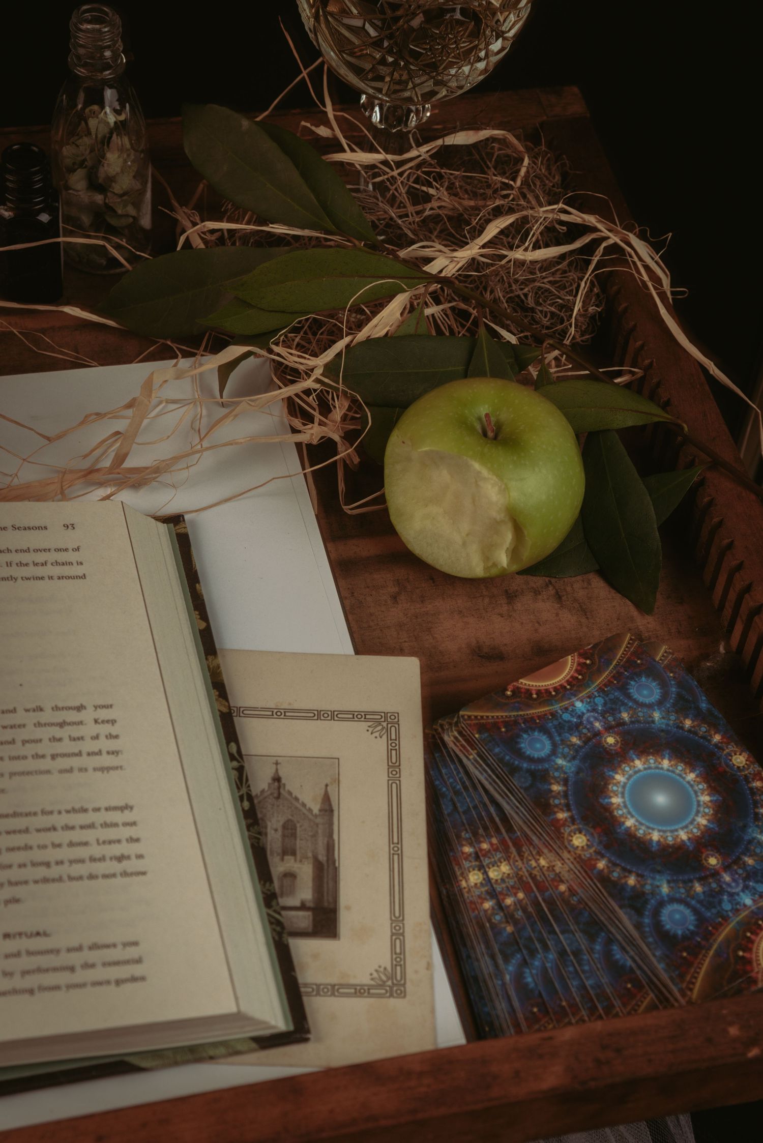 Open books with mystical illustrations and green apples arranged on a dark wooden surface.