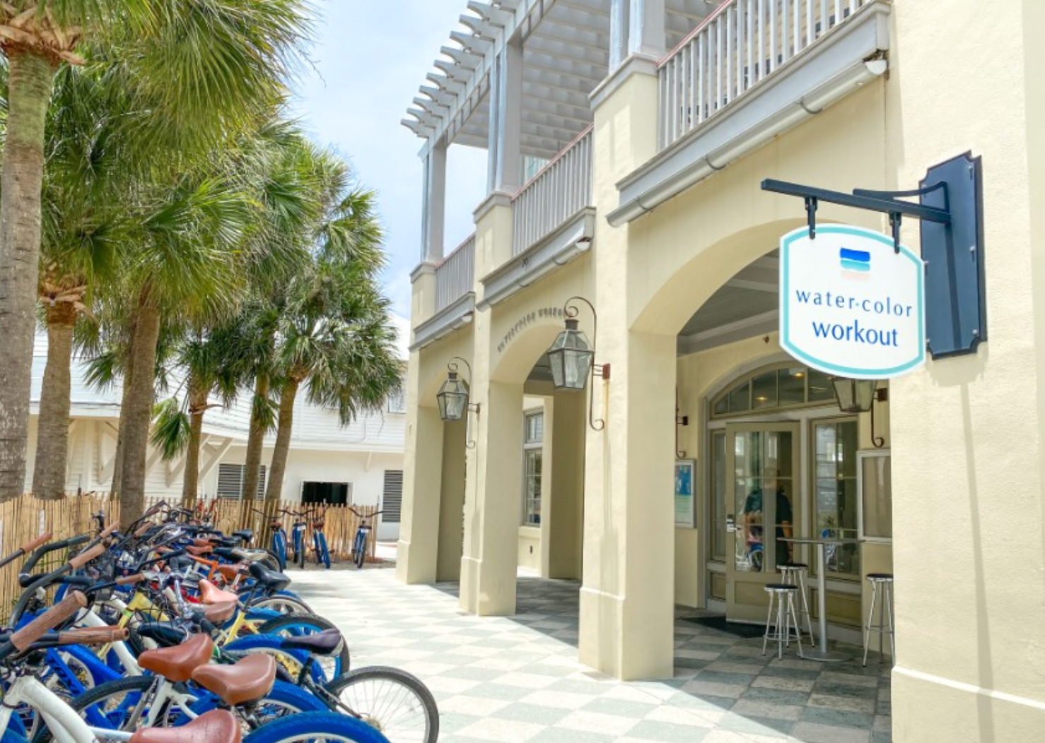 Row of colorful beach cruiser bicycles parked under white columns along a beachfront walkway.