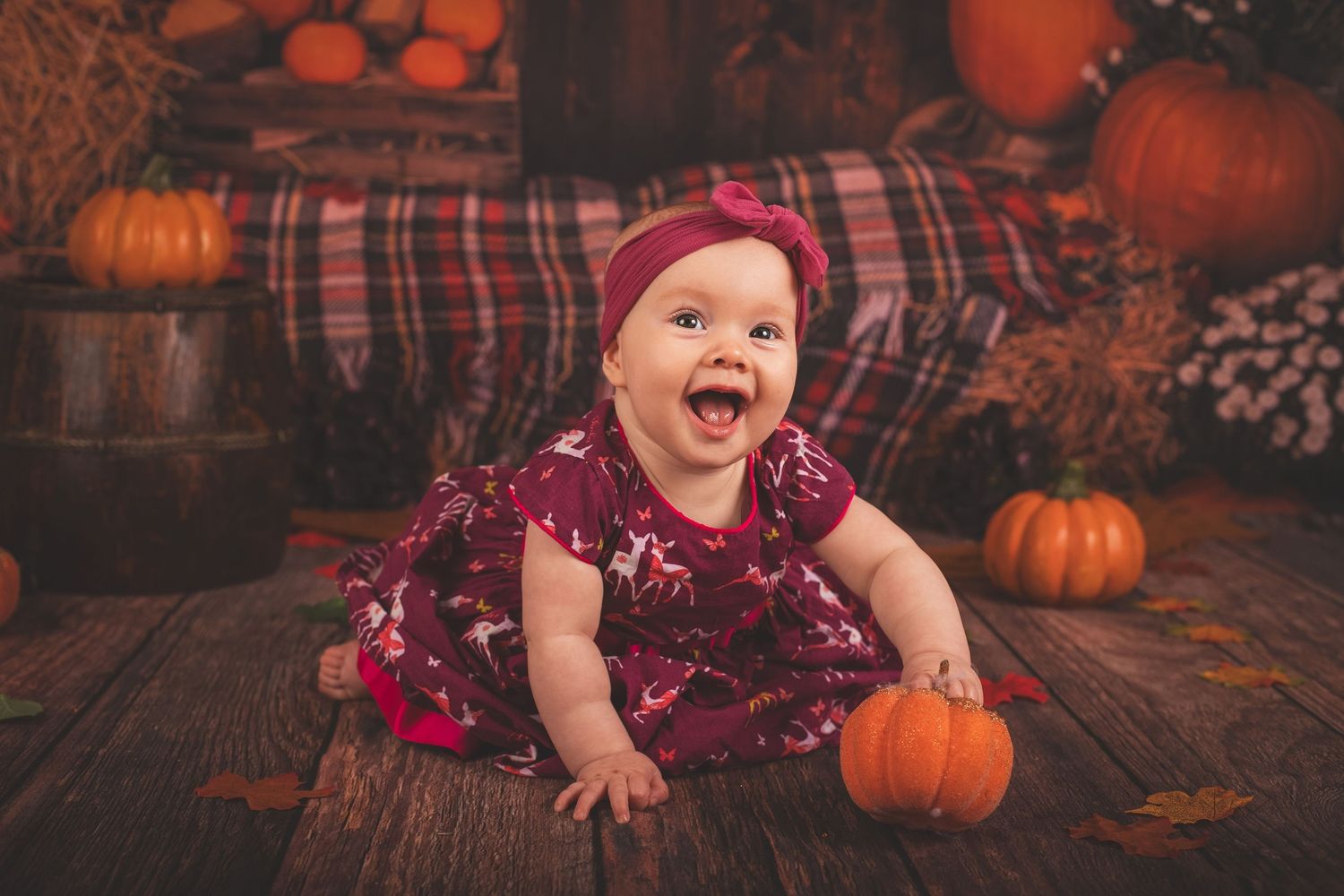 A joyful baby in a burgundy dress sits among autumn pumpkins and plaid decorations on a rustic wooden floor.