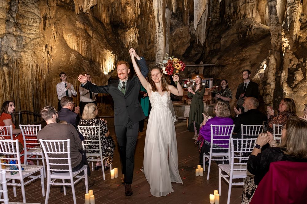 A couple celebrates during their wedding ceremony inside a dramatic cave setting with white chairs and natural stone formations.