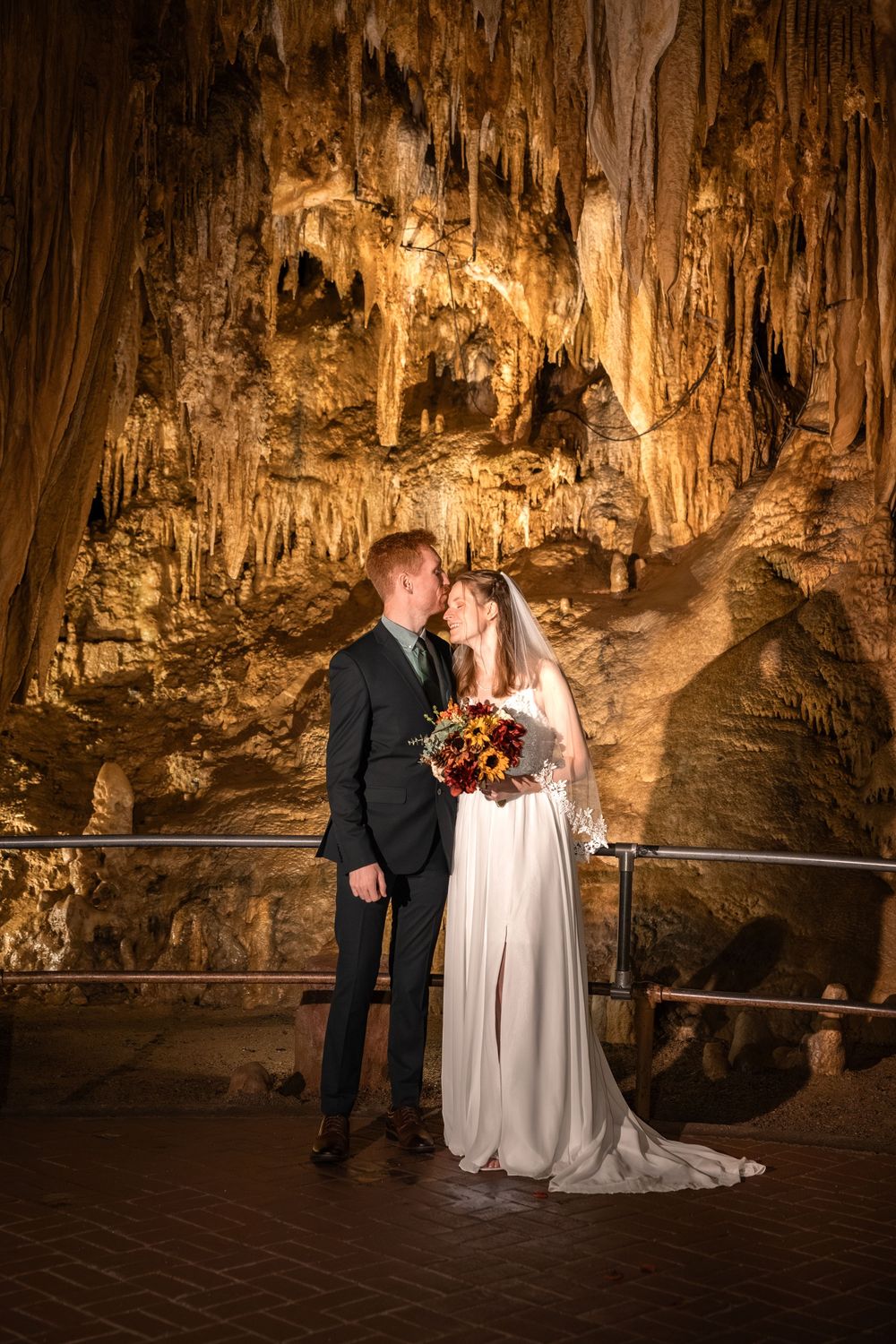 A wedding couple stands on a walkway inside a dramatic cave formation with illuminated stalactites and stalagmites.