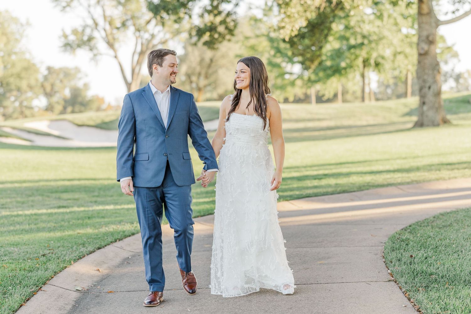 A couple walks hand in hand along a sunlit path in a lush green park during a romantic outdoor photoshoot.