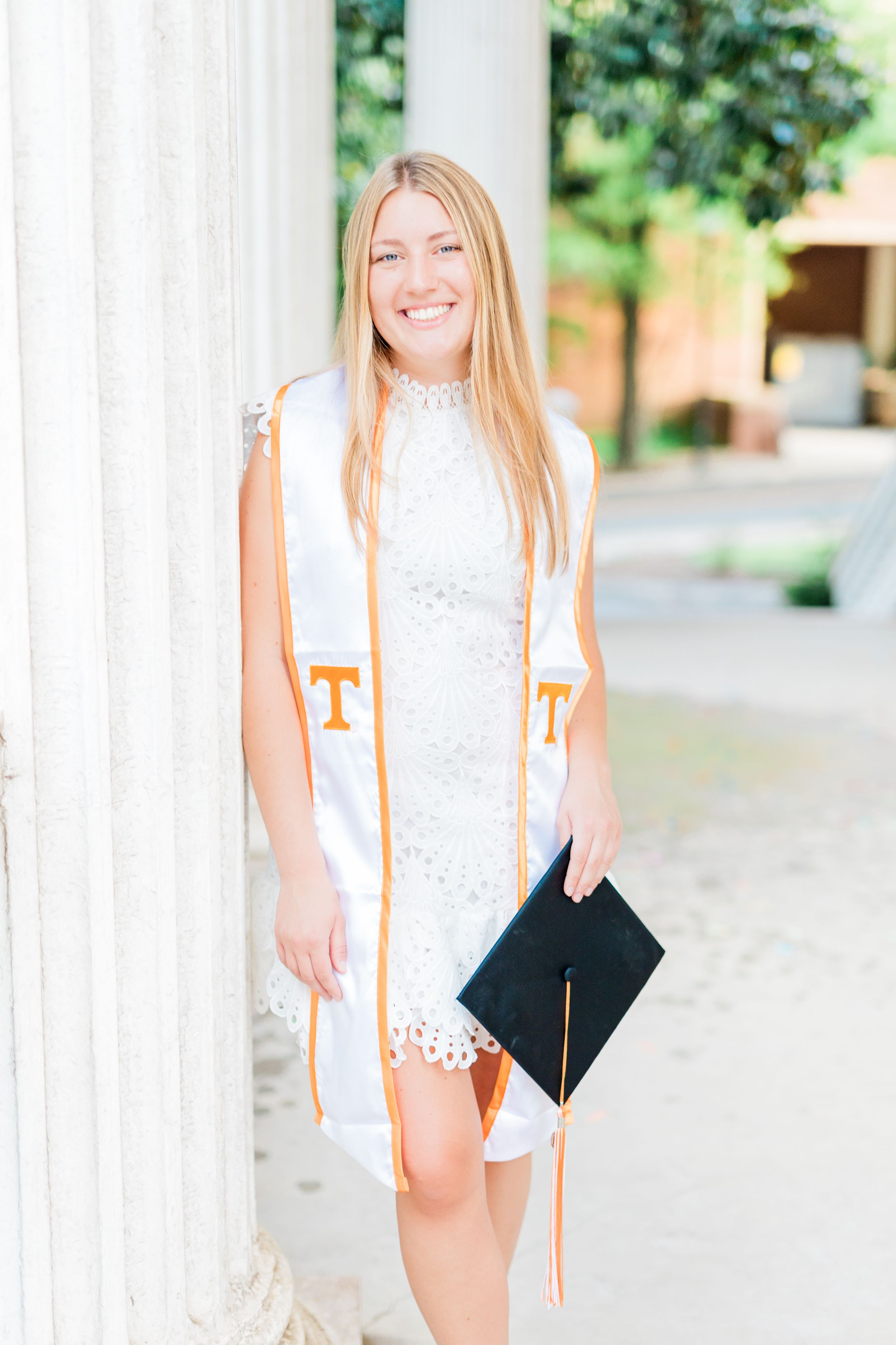 A graduate poses in white lace dress with orange graduation stoles and cap for outdoor senior portraits.