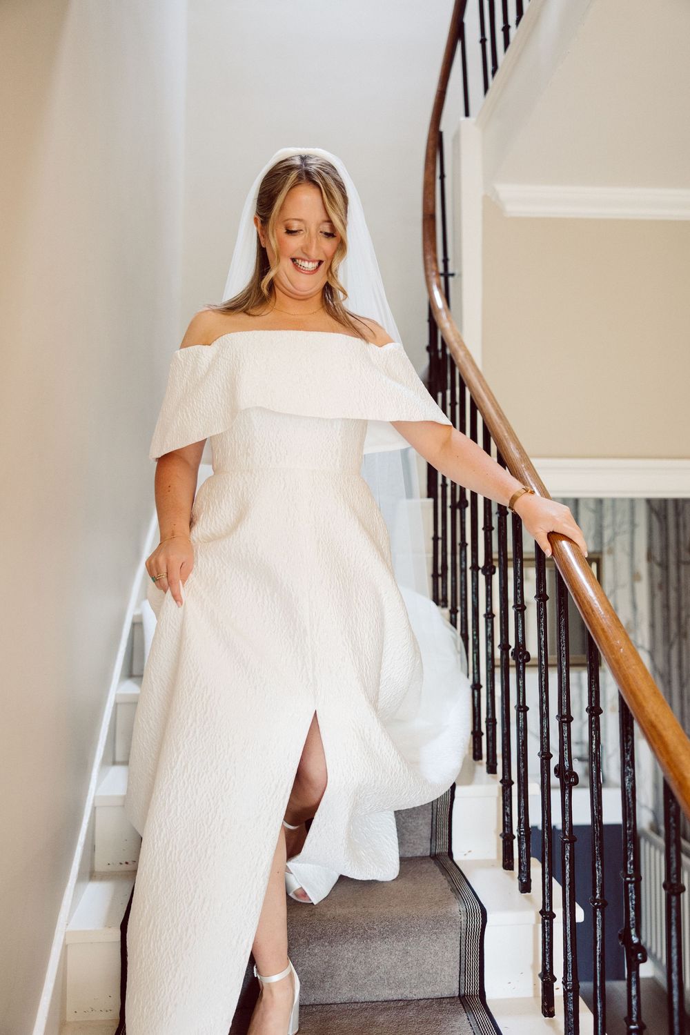 A bride in an off-shoulder white wedding dress descends a staircase while holding the railing and smiling joyfully.