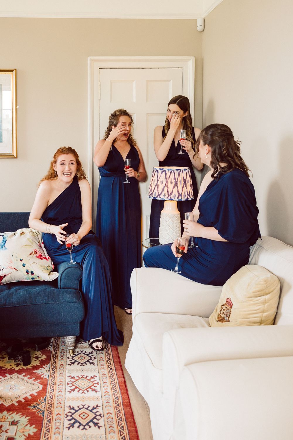 Two friends in navy blue dresses share a joyful moment on a white couch with a decorative rug below.