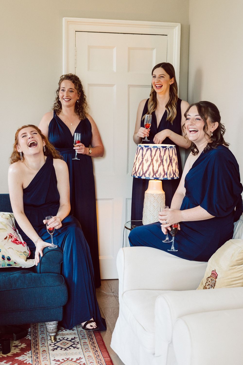 A group of friends in navy blue dresses share laughter and wine while relaxing on couches in a living room setting.