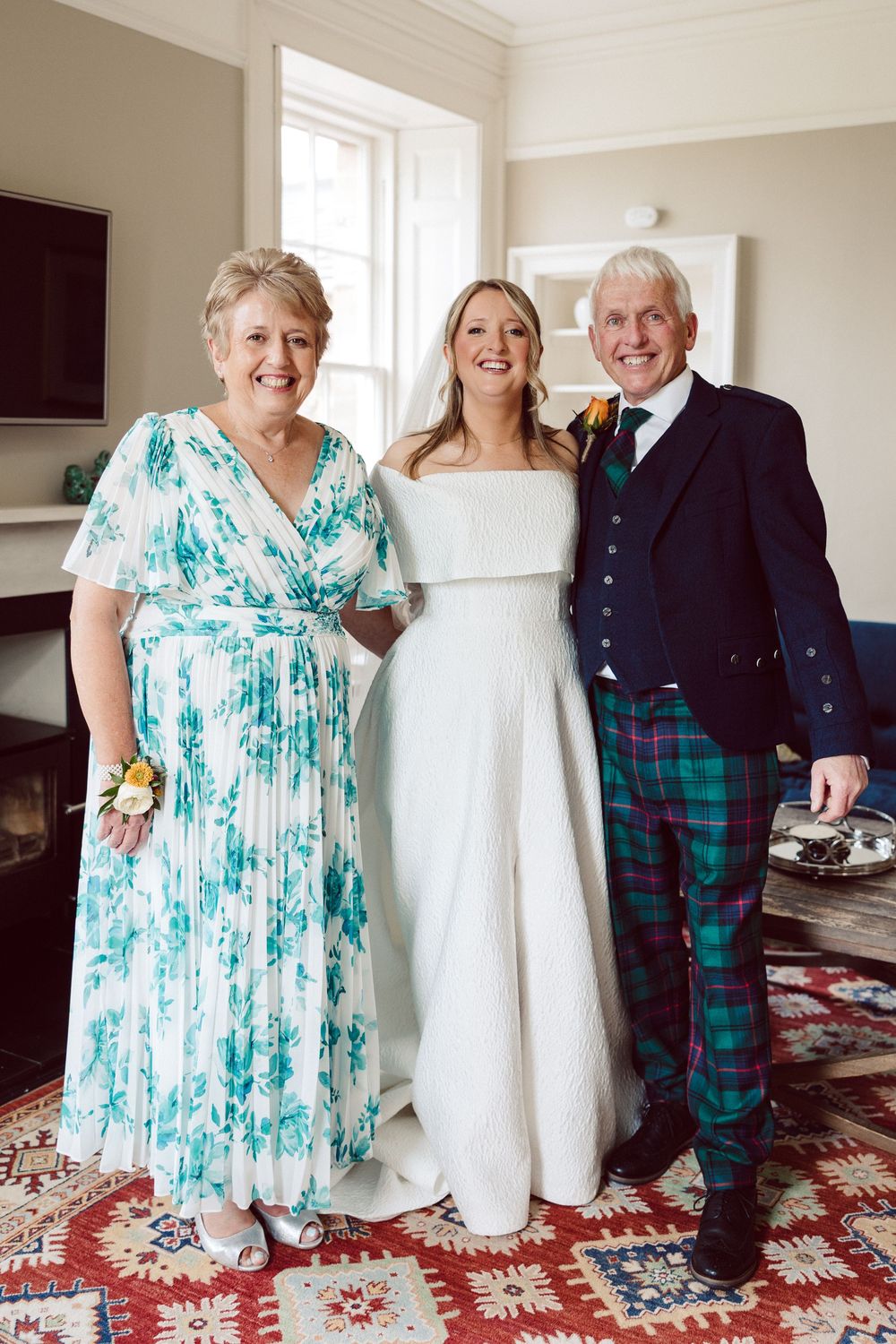 A wedding party poses together on a patterned carpet, featuring traditional Scottish attire and formal wear.