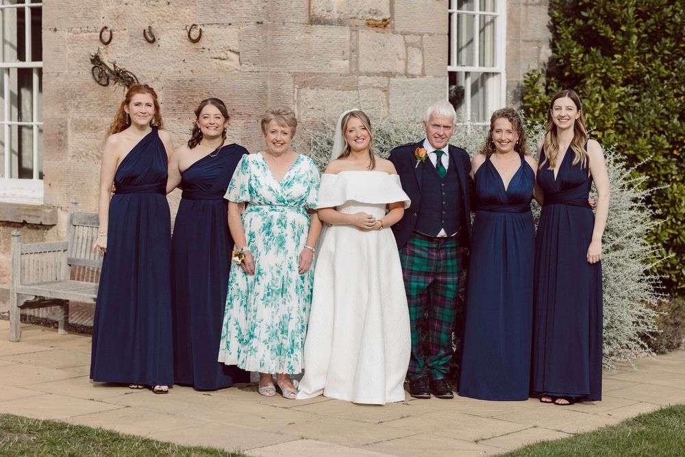 Wedding party in navy blue dresses and Scottish tartan kilt poses together outside stone building.
