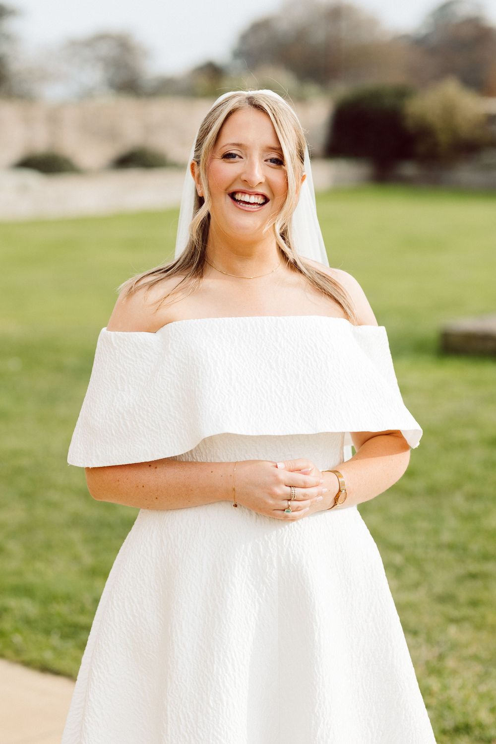 A flowing off-shoulder white dress is showcased in an outdoor setting with green grass and trees.