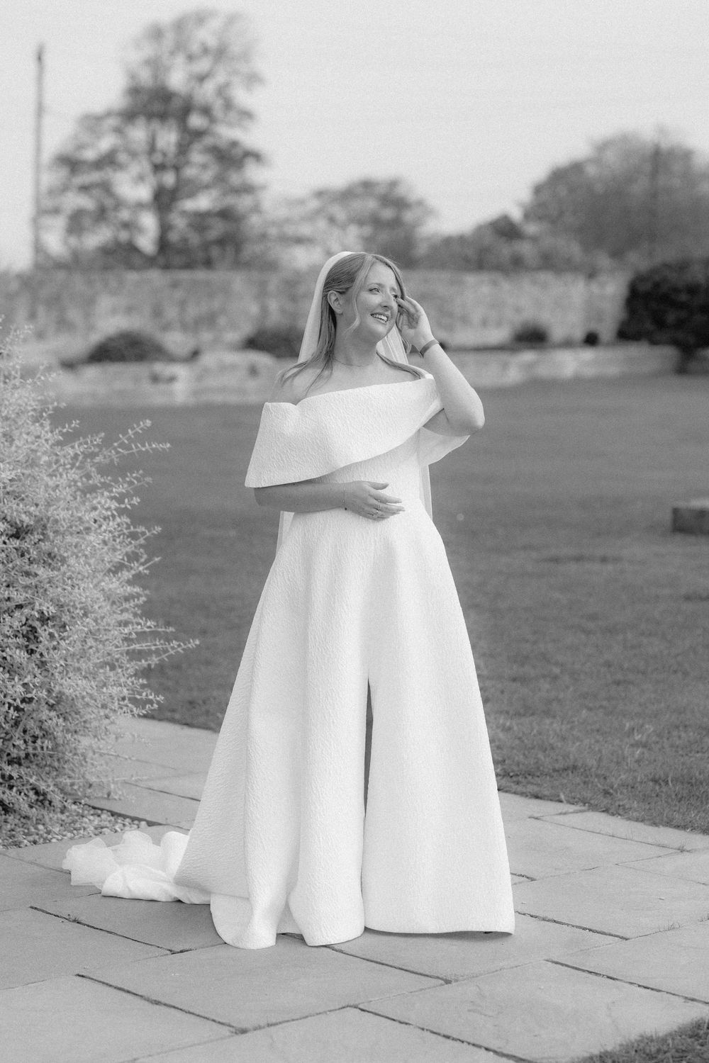 A black and white photo shows an elegant off-shoulder white dress against a natural outdoor backdrop.