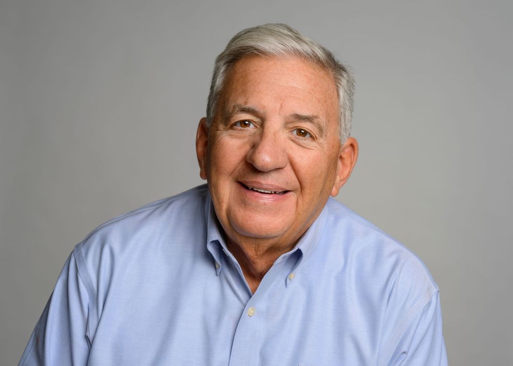 Smiling older man with gray hair wearing a light blue shirt poses for a portrait.