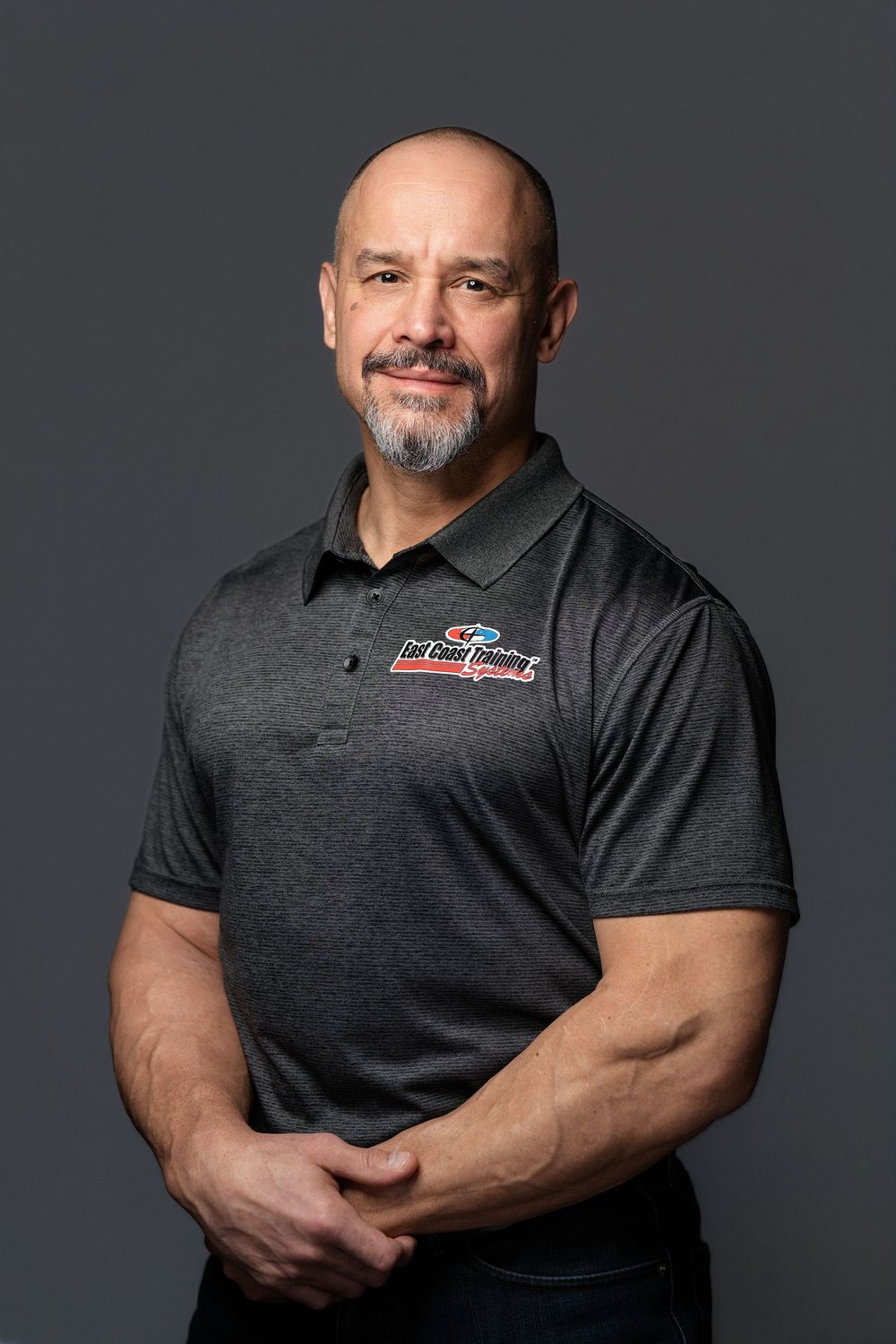 Muscular bald man with goatee wearing a gray polo shirt poses for a professional portrait.
