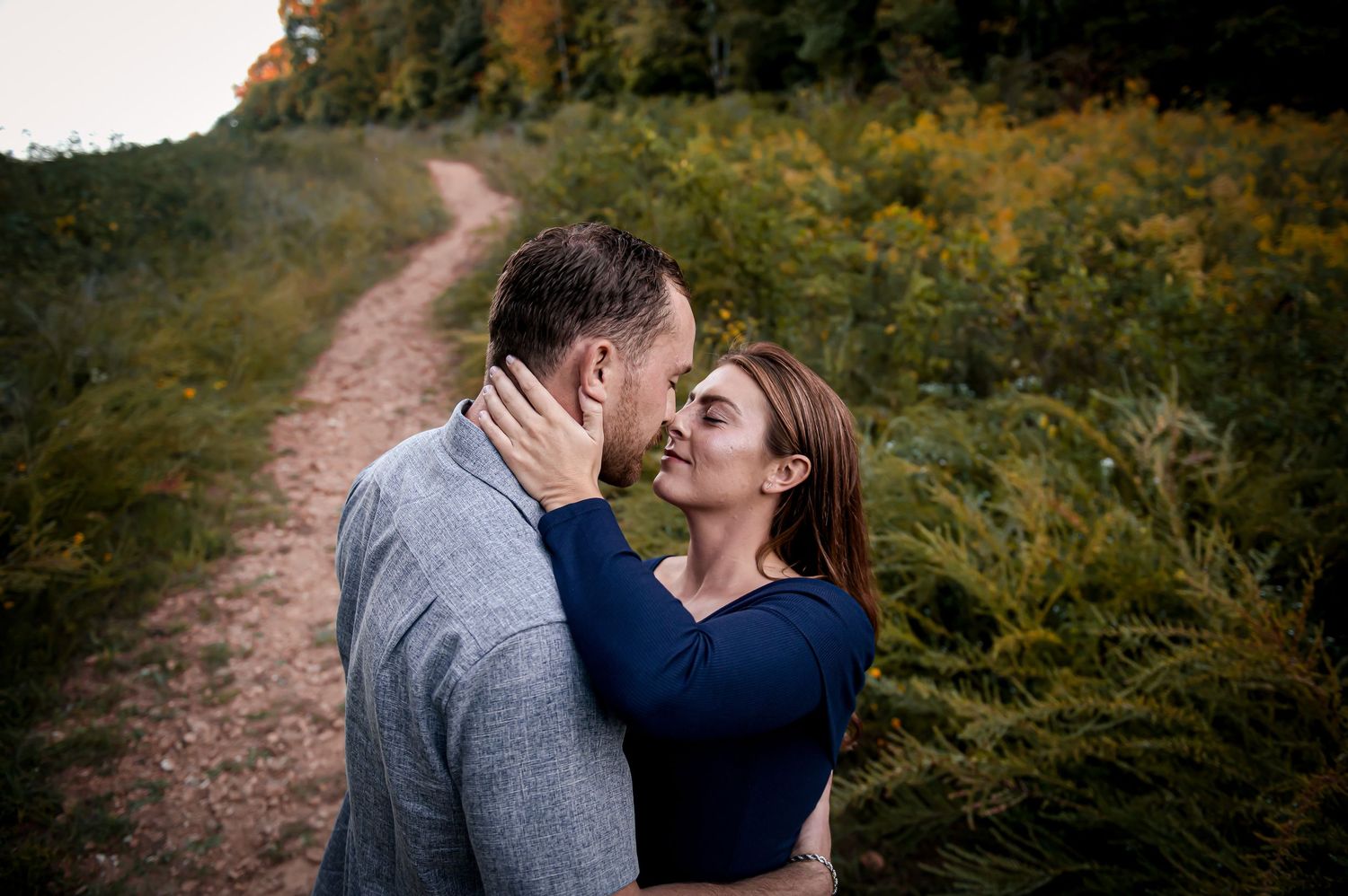 Two people share an intimate moment on a winding dirt path surrounded by lush green foliage.