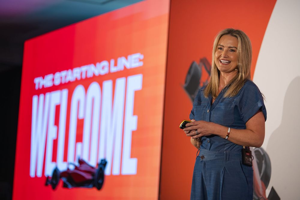 Woman smiling on stage at a welcome presentation with a red backdrop reading The Starting Line: Welcome.