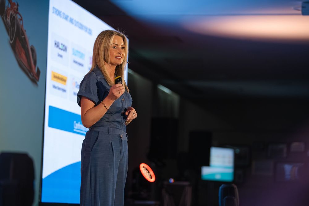 Professional female speaker presenting on stage with microphone, blue backdrop screen at corporate conference event.
