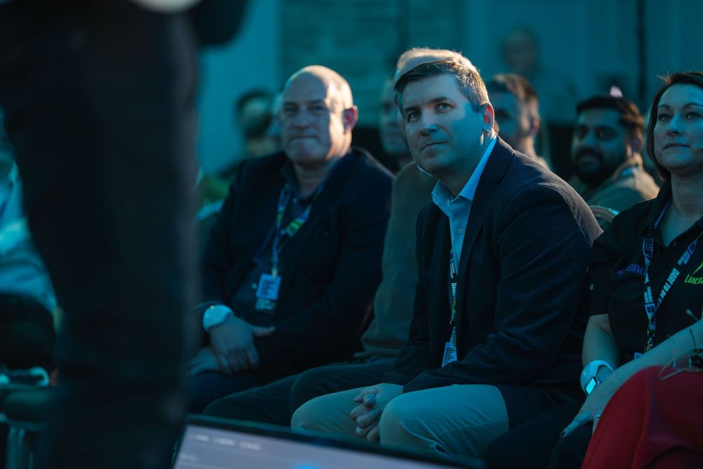 Audience members watch intently in a dark auditorium with blue lighting casting shadows across the crowd.