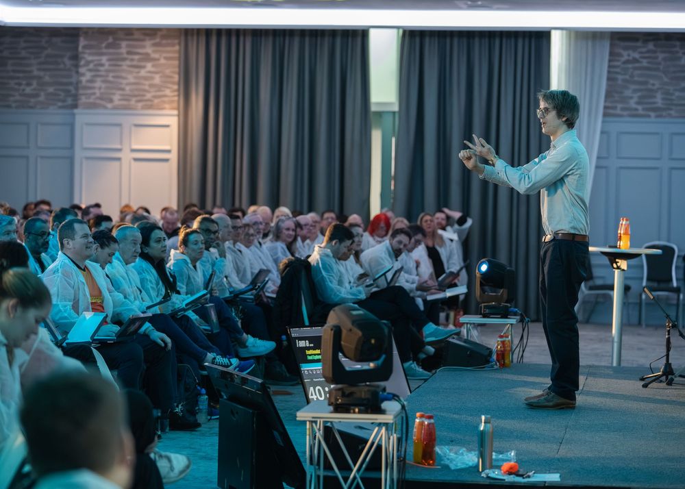 Speaker presenting to a packed audience in a conference room with blue ambient lighting and dark curtains in the background.
