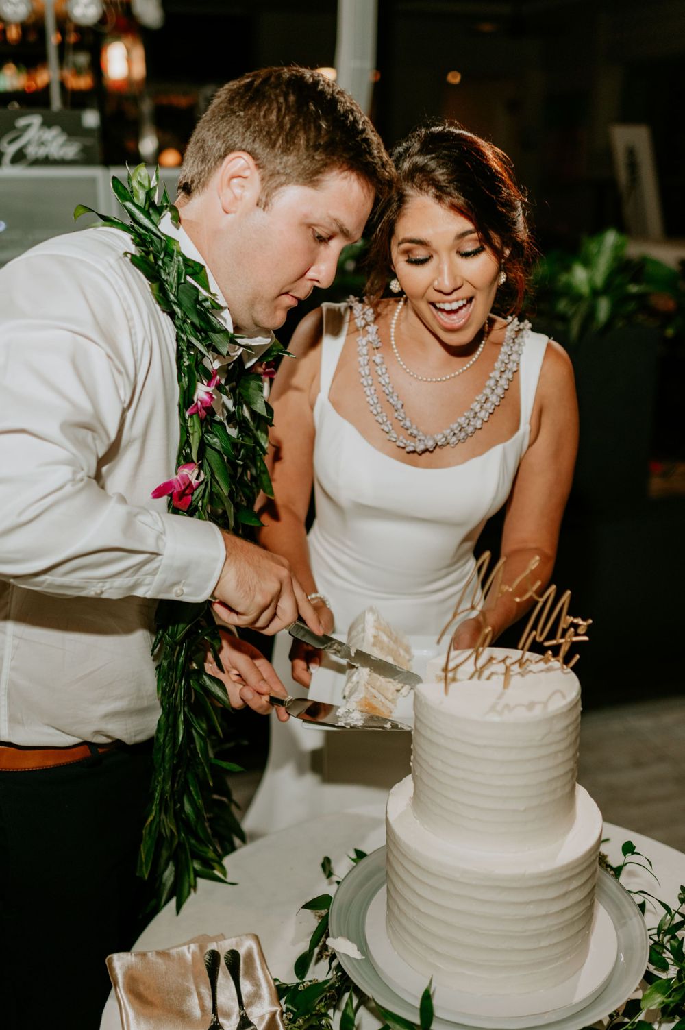 Couple cutting their elegant white tiered wedding cake decorated with greenery.