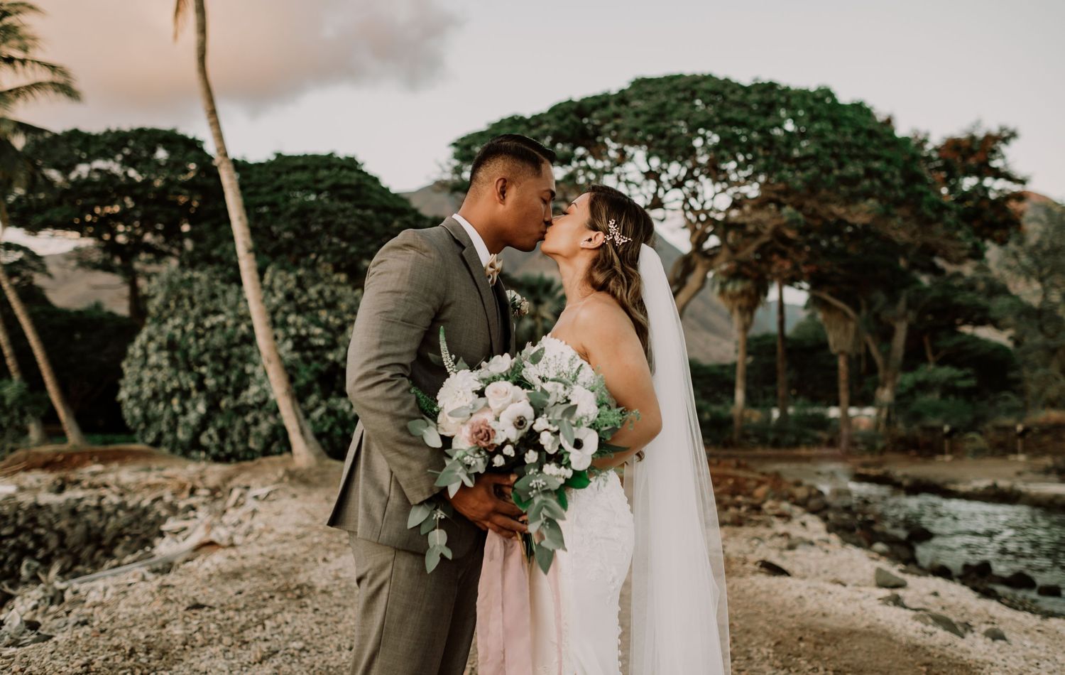 A couple shares an intimate moment during their beach wedding photoshoot with lush greenery backdrop.