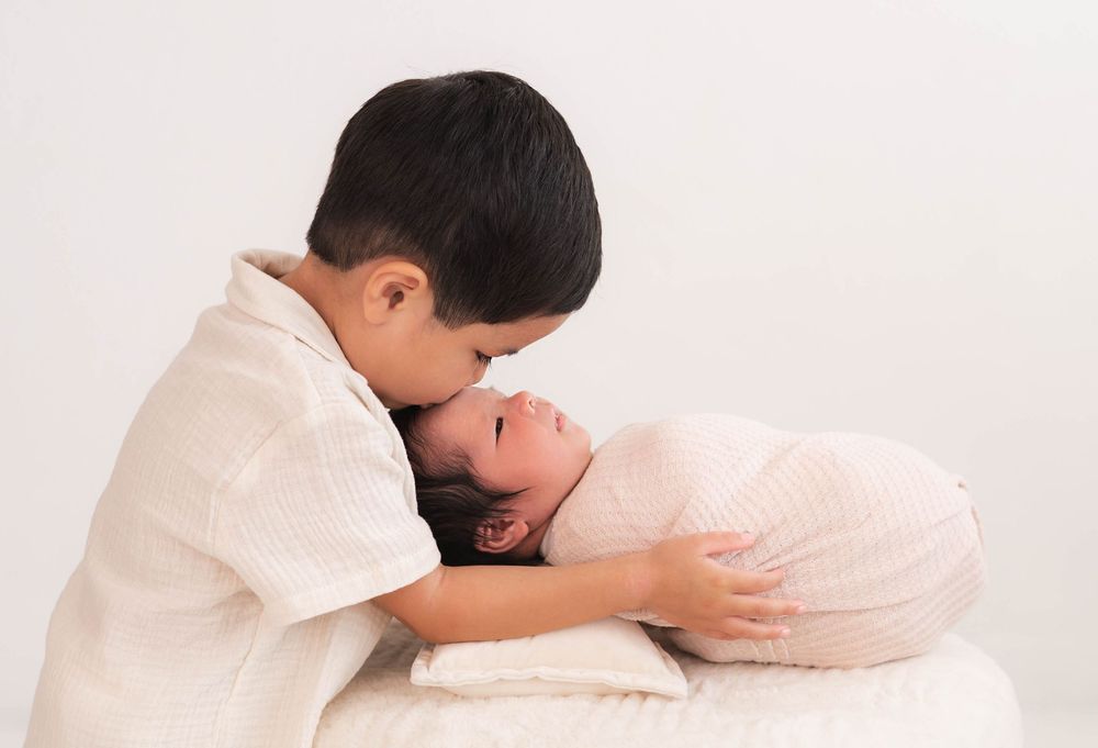 Young boy gently kissing newborn baby sibling wrapped in pink swaddle on white background.