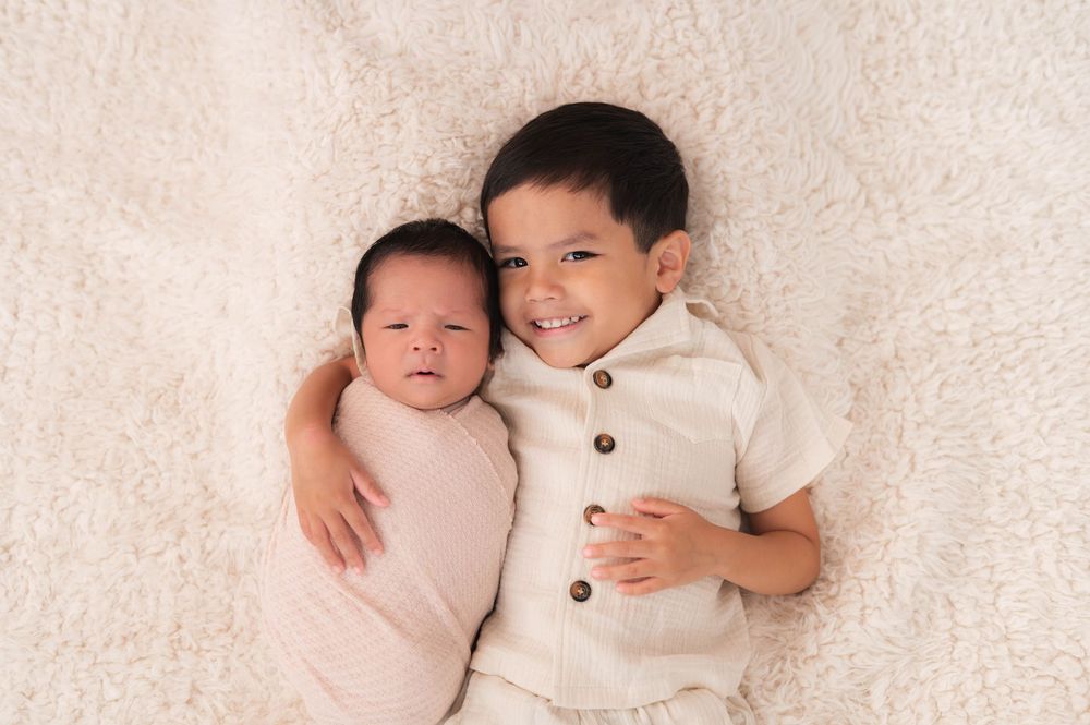 Newborn baby swaddled in pink wrap lying next to smiling toddler boy on a cream fluffy blanket.