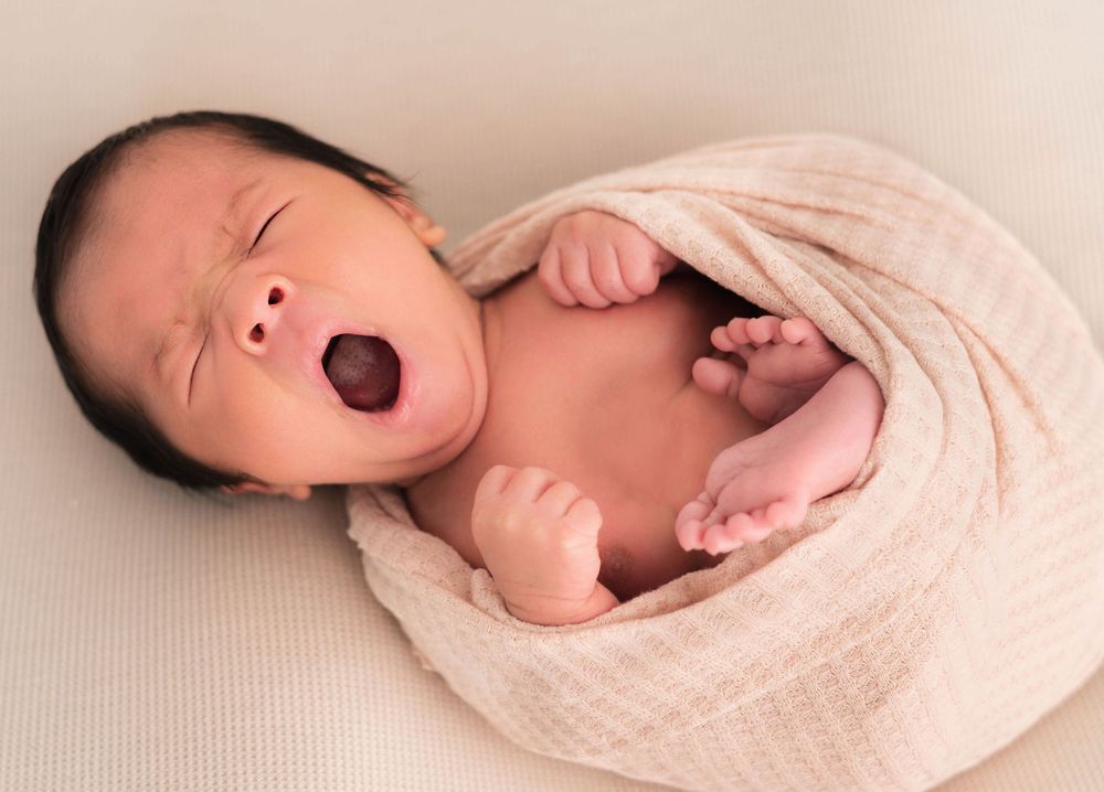 Newborn baby yawning while wrapped in a soft pink swaddle blanket, with tiny fists raised in a peaceful pose.