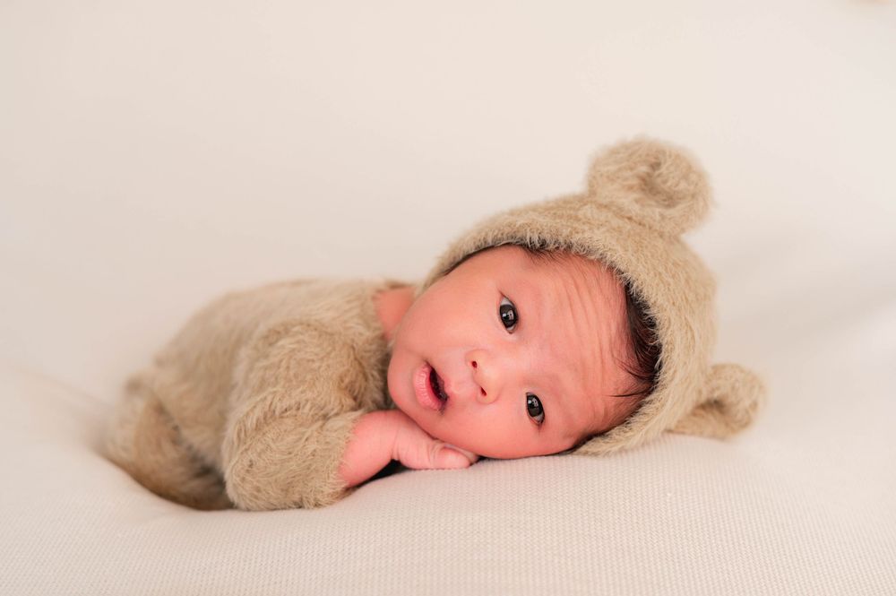 Newborn baby in a cozy beige knit bear outfit with ears, lying on a soft white blanket.