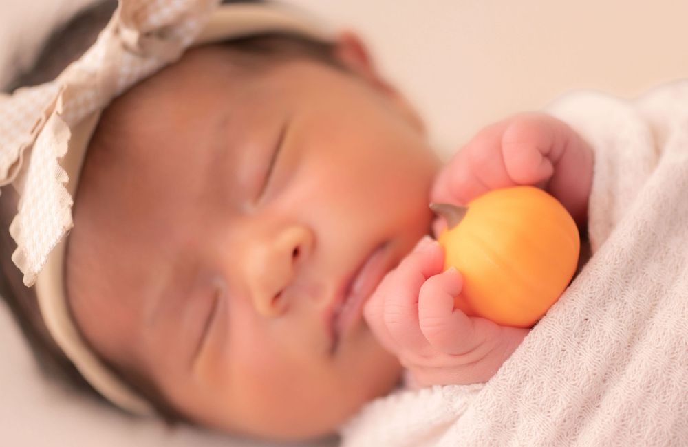 Sleeping newborn baby girl wearing a bow headband, wrapped in a pink blanket, holding a small orange fruit.