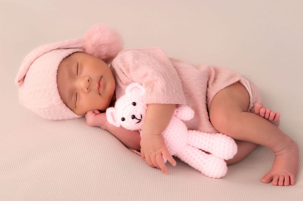 Newborn baby girl sleeping peacefully in pink knit outfit and hat, cuddling a pink crocheted teddy bear.
