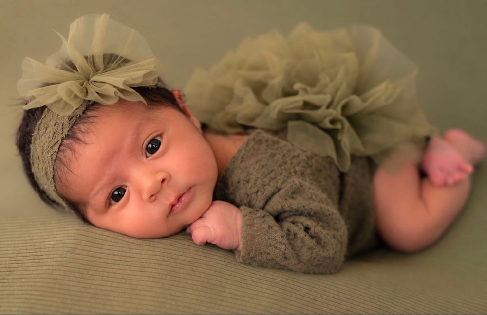 Newborn baby girl in olive green tutu skirt and floral headband lying on a green backdrop.
