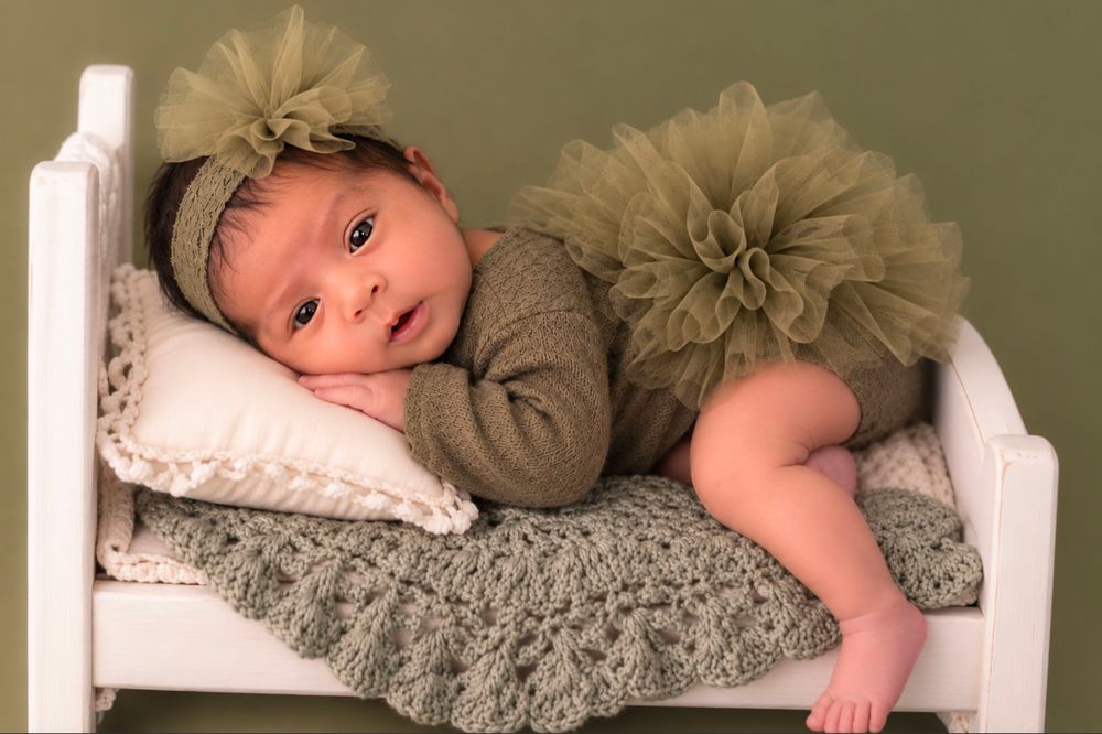 Newborn baby in olive green tutu and headband posed on a tiny white bed with knit blanket.