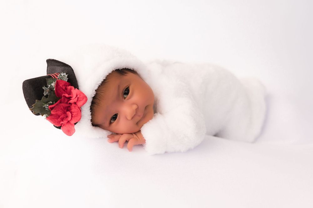 Newborn baby in white fluffy outfit with black hat and red flowers, posed on white background.