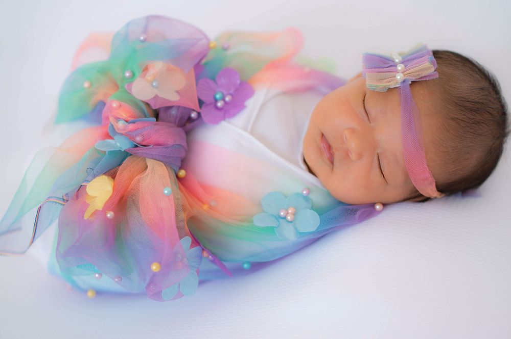 Sleeping newborn baby wrapped in colorful rainbow organza fabric with pearl details and pink bow headband.