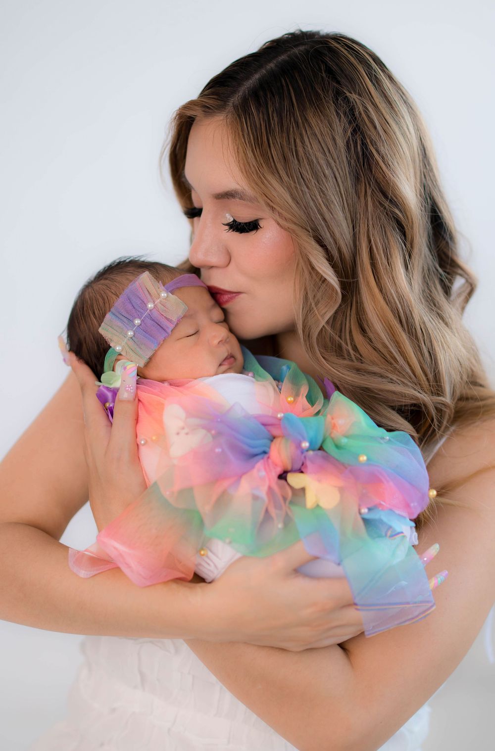 A young mother with long blonde hair gently kisses her sleeping newborn dressed in a colorful rainbow tutu outfit.