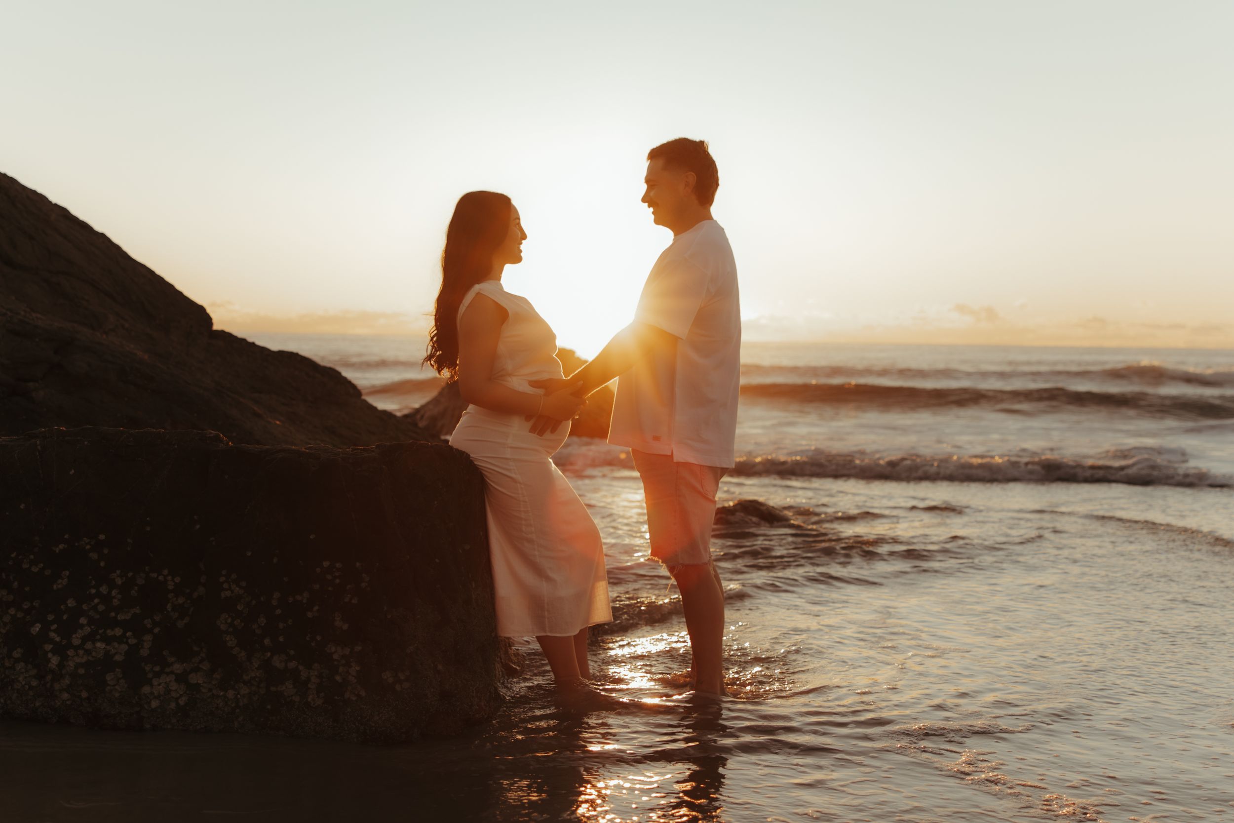 Silhouetted couple embraces in shallow ocean water during golden sunset at beach.