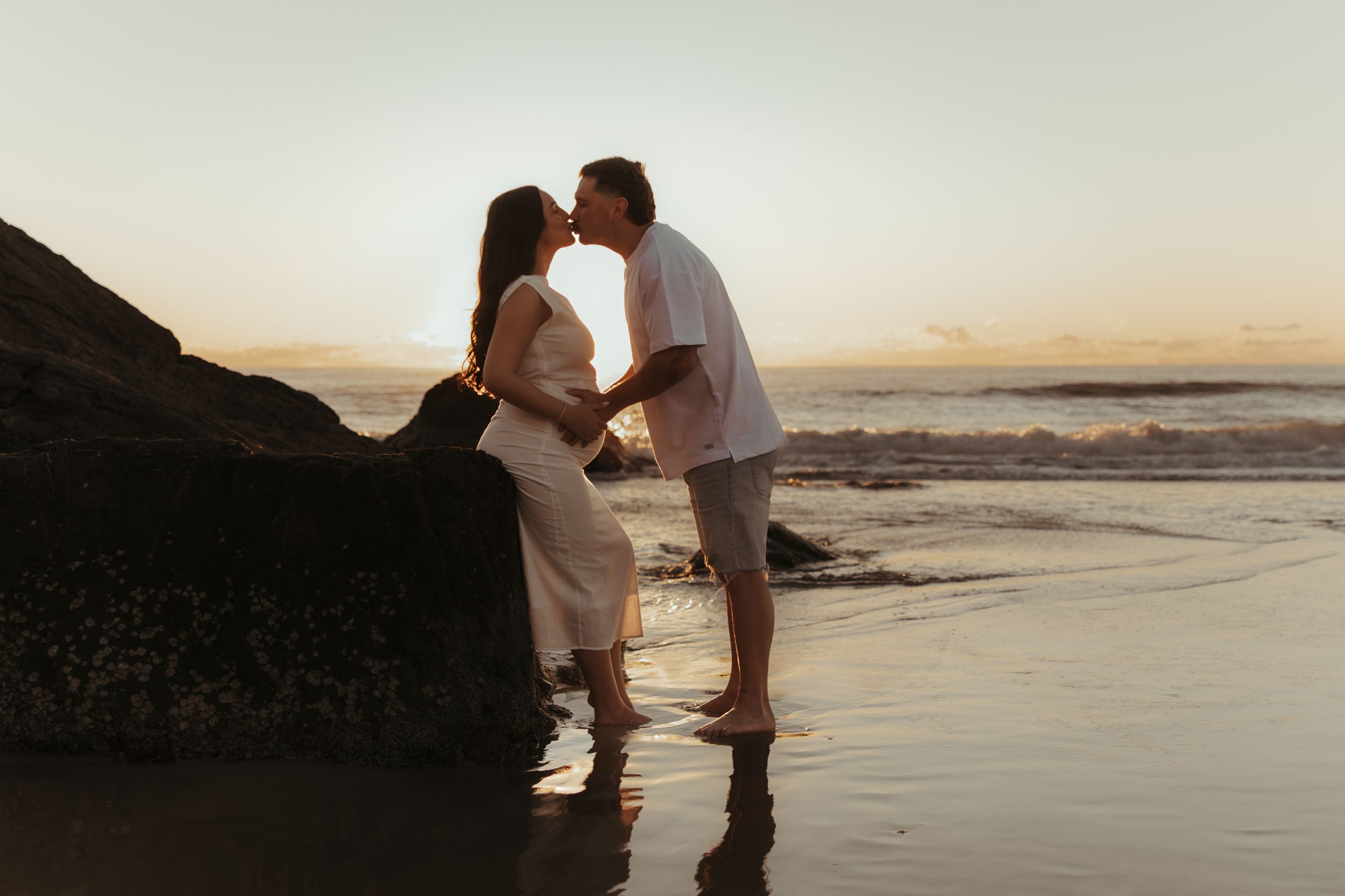 Two silhouettes embrace on a sandy beach during sunset with ocean waves in the background.