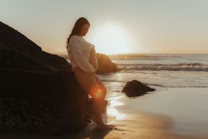Silhouetted couple sits on rocky beach watching golden sunset over ocean waves.